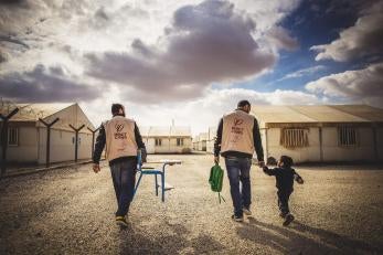 Two prosper global team members and a small boy walking; one man is carrying a chair and the other man is carrying the boy's backpack and walking hand-in-hand with the boy.