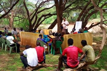 Kenyan community members meet in the outdoors under trees.