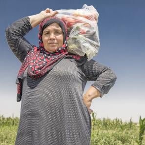 A woman in syria carries a plastic bag filled with vegetables on her shoulder